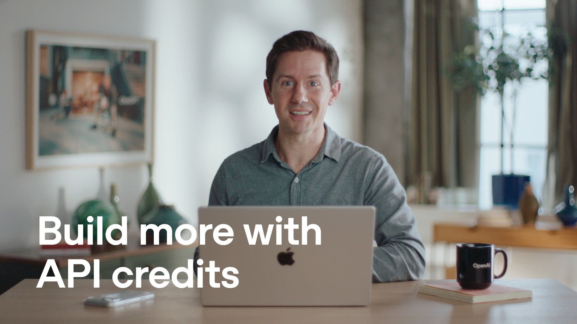 A still from OpenAI's announcement video: a man smiling at a laptop on a wooden table next to an OpenAI mug, with the words "Build more with API credits" in white.