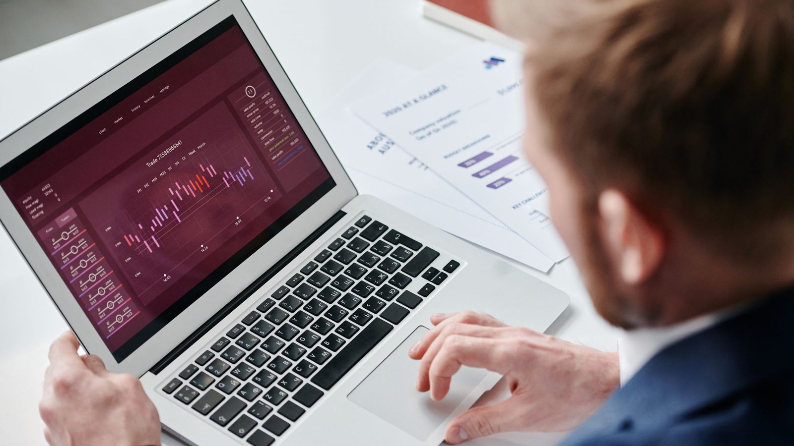 A person in a suit, seen from behind, reviewing an analytics dashboard on a laptop next to printed reports.